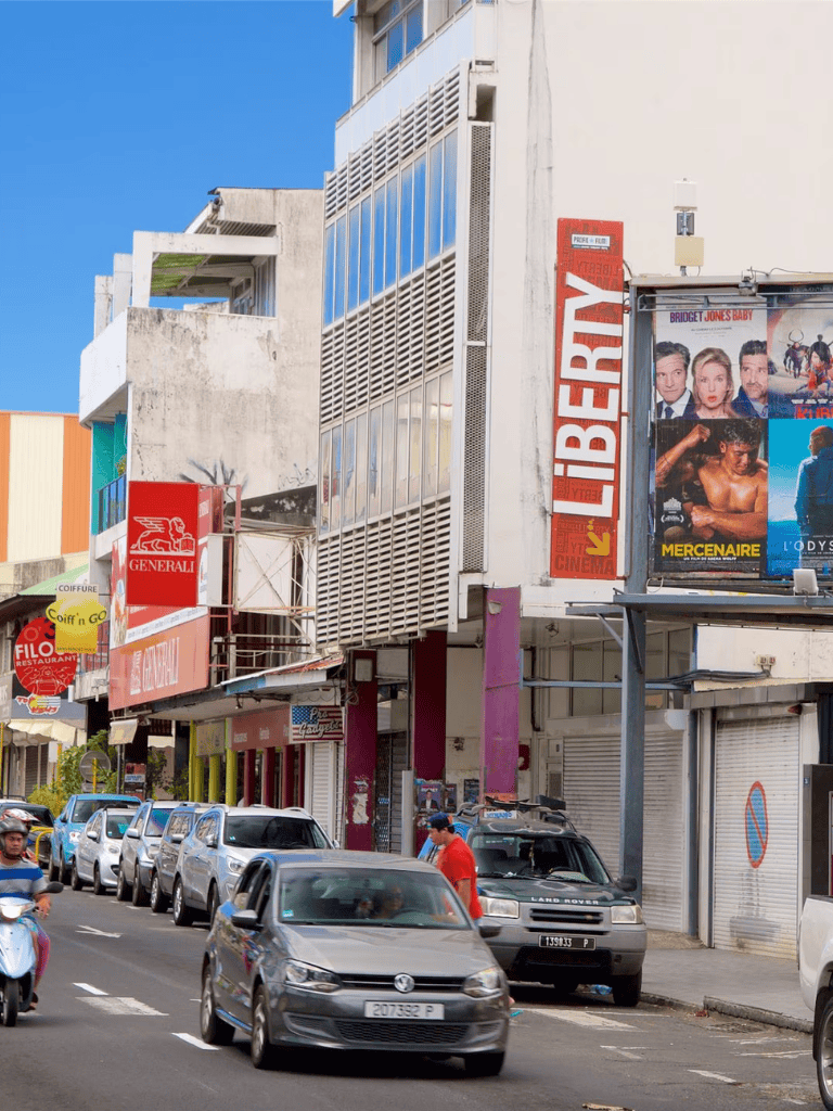 Busy city street with multiple cars, pedestrians, and colorful signage, including a cinema and various storefronts.