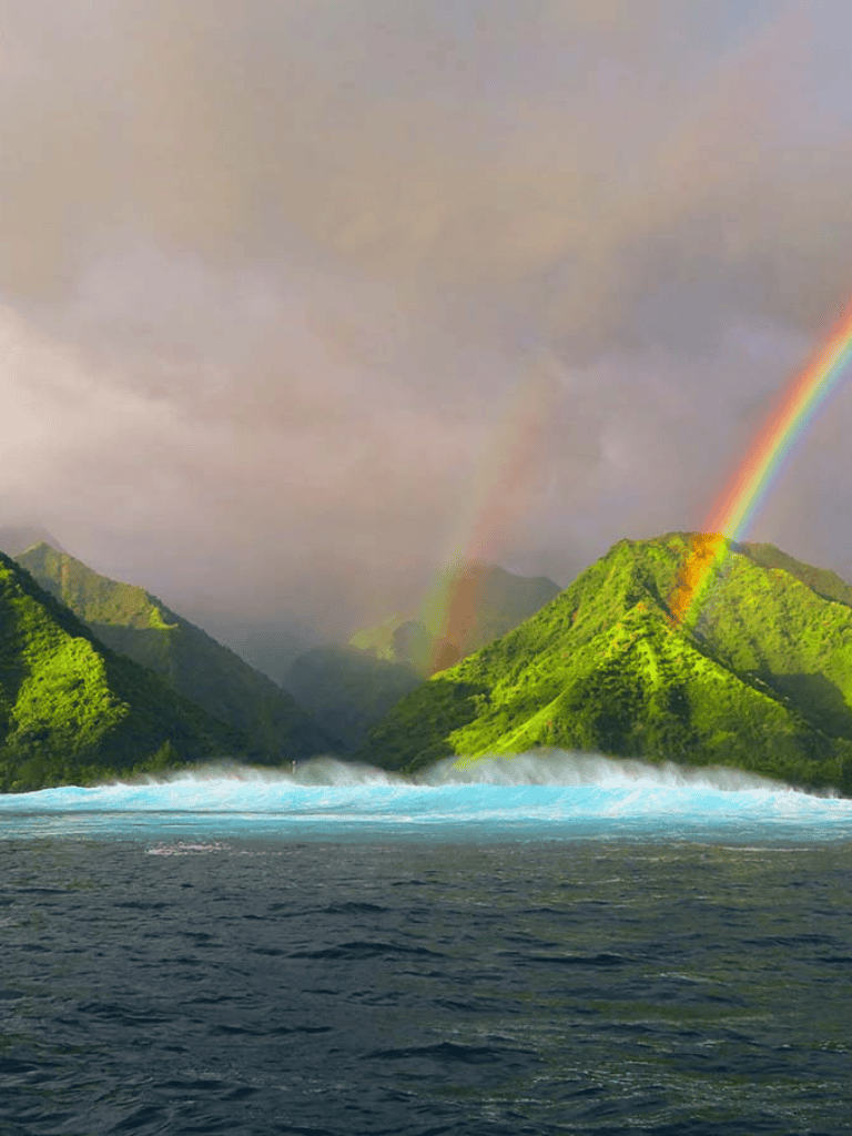 Vivid rainbow over lush green mountains and ocean waves, stunning natural scenery and travel destination.