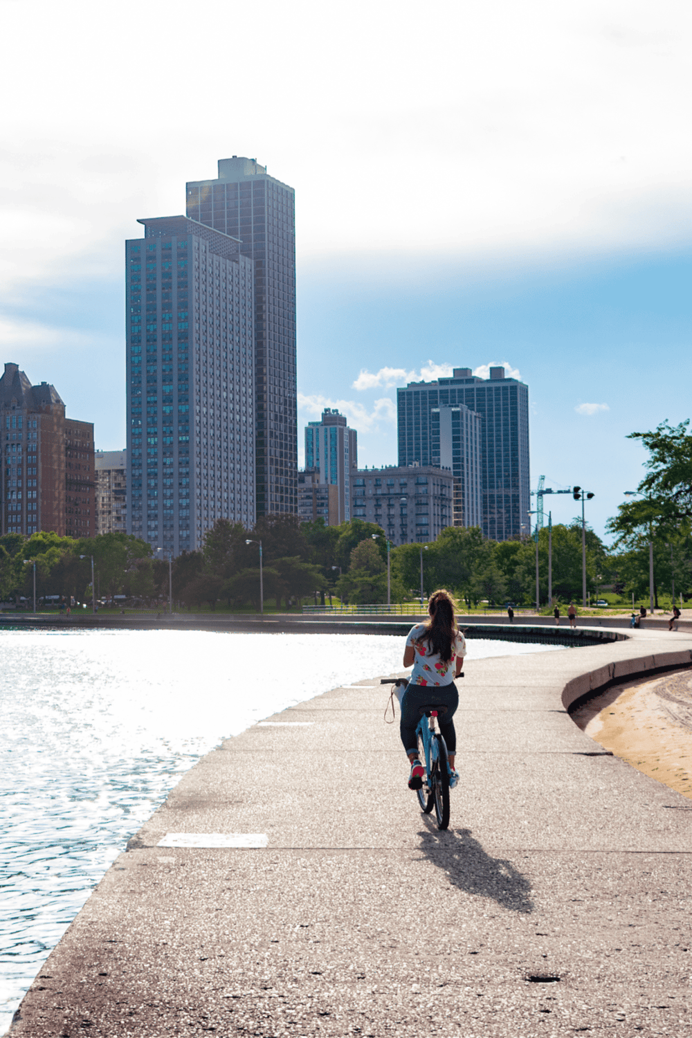 Bicycle rider along city waterfront with skyscrapers, urban park, and clear sky in the background.