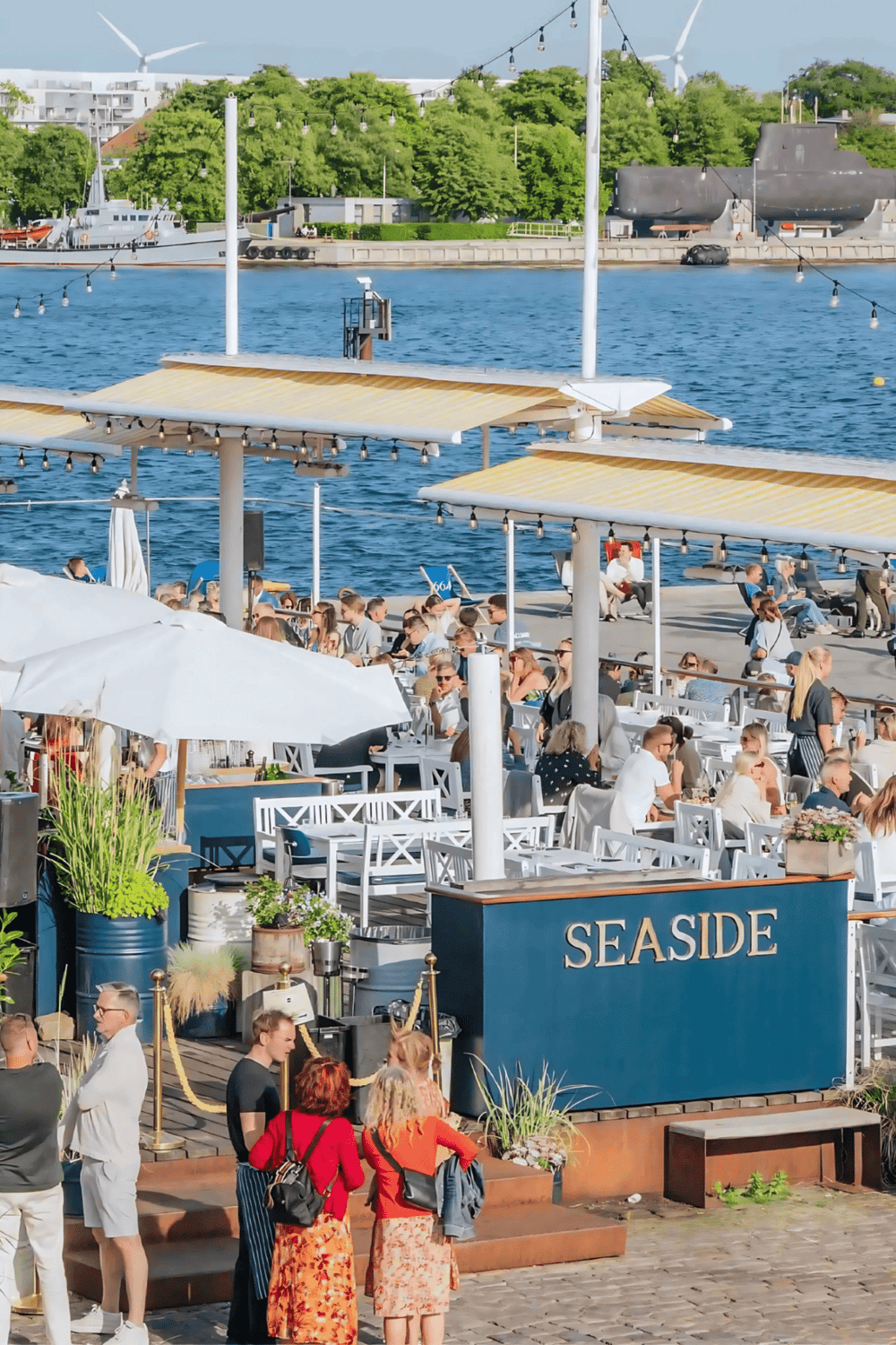 Outdoors waterfront dining at Seaside restaurant, bustling crowd enjoying scenic views and ocean breeze.