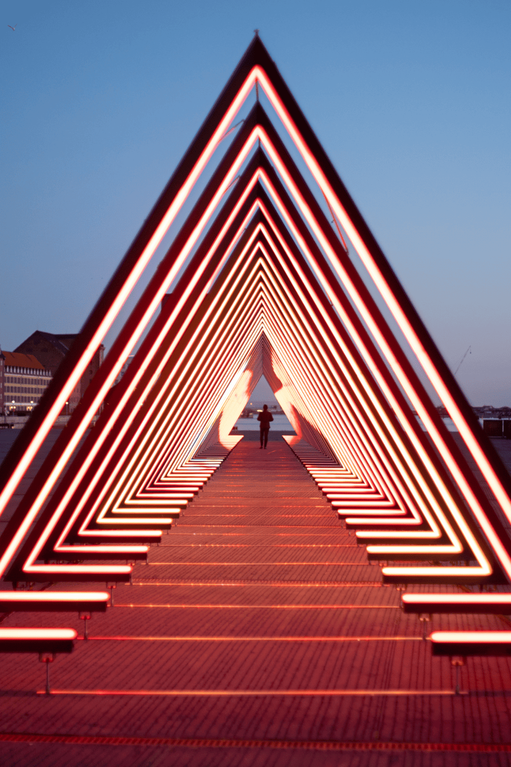 Bright neon triangular light installation on a pier at dusk, perfect for travel and attraction keywords.