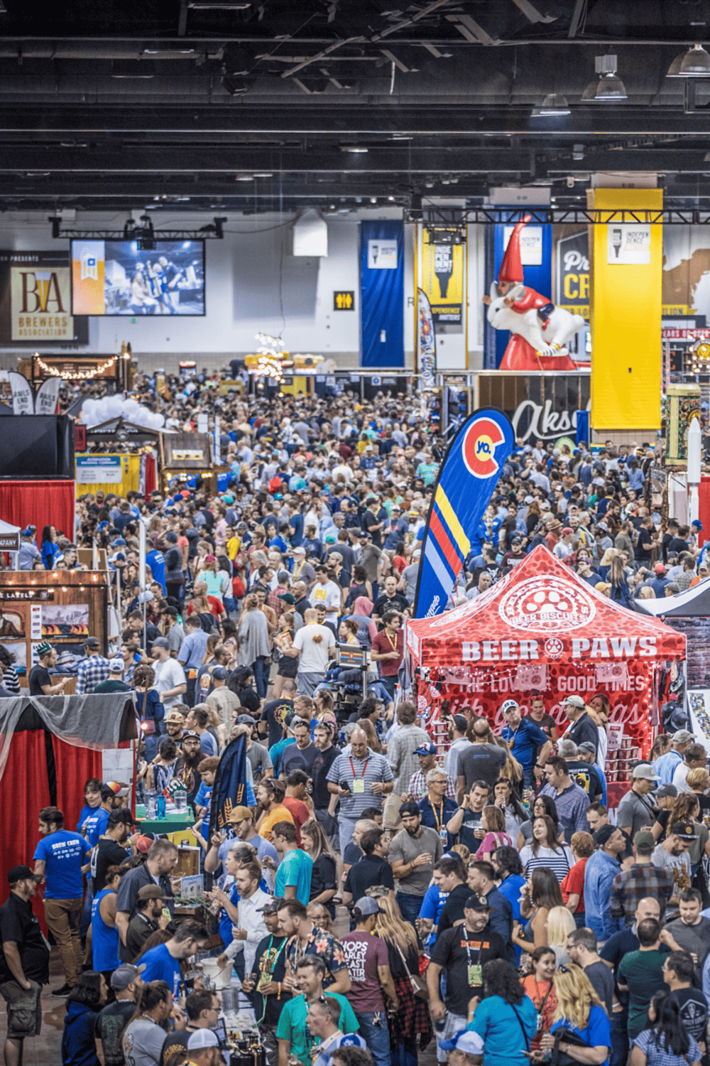Crowd at a large trade show or convention with beer and food vendors, vibrant banners, and indoor entertainment.