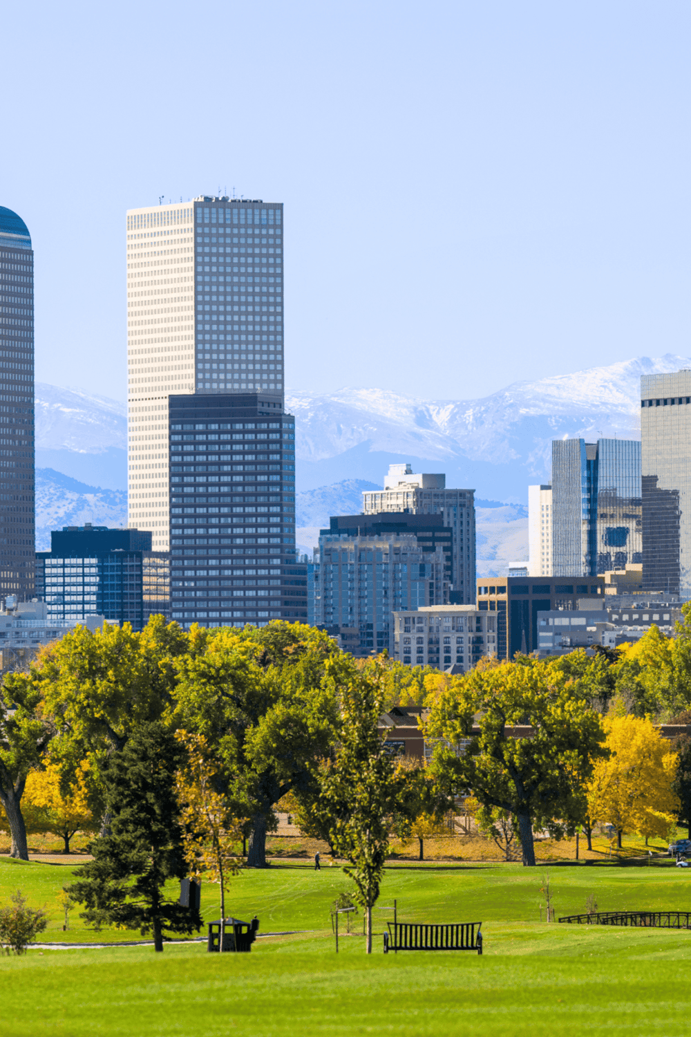 City skyline with tall modern buildings and mountain backdrop, featuring a lush park in the foreground.