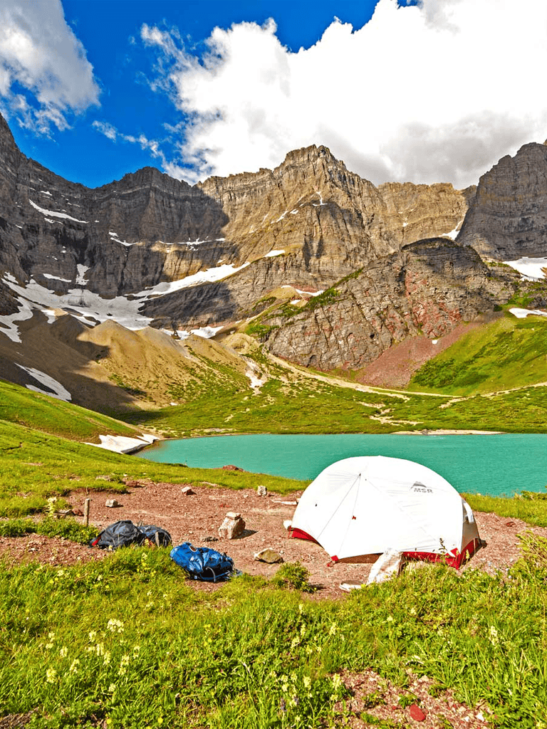Vibrant mountain landscape with lake, camping tent, and backpacks in lush green valley under blue sky.
