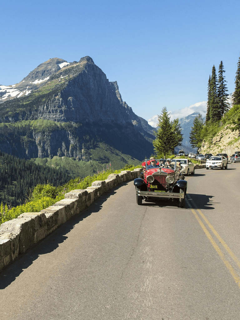 Vintage car driving scenic mountain road with lush greenery and towering peaks.