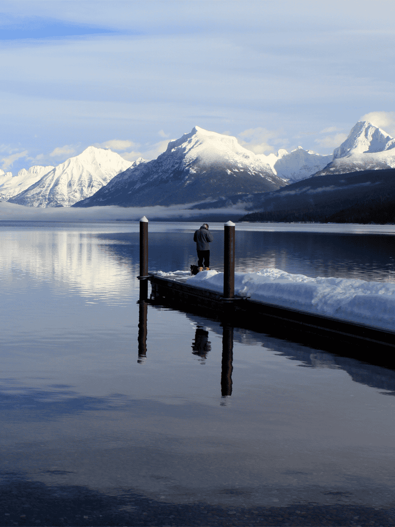 Snow-capped mountains reflecting on calm lake with a person standing on dock in winter.