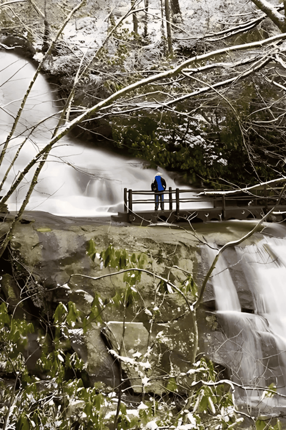 Snow-covered forest waterfall with hikers on a wooden bridge, scenic winter outdoor adventure.