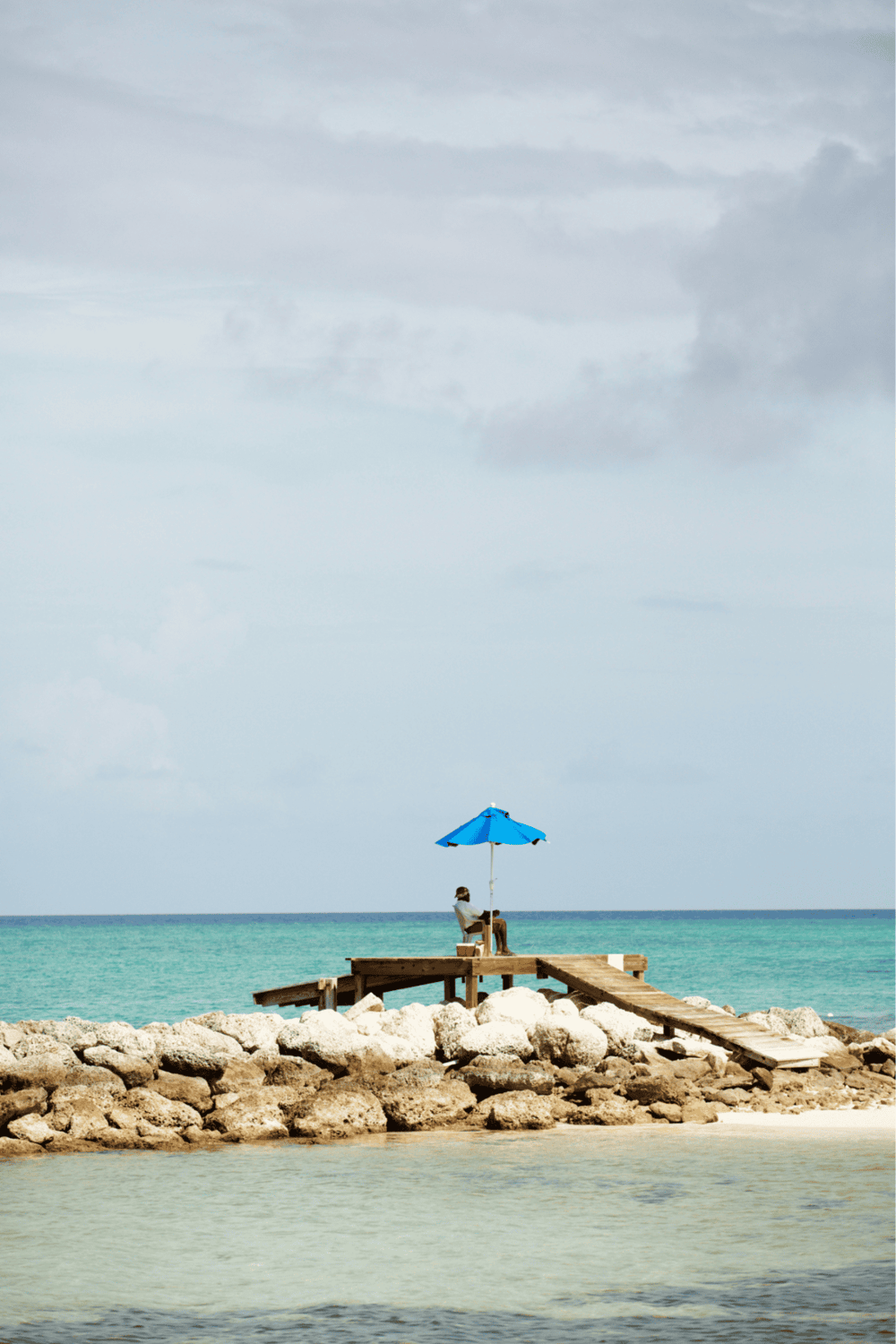 Tranquil beach scene with a person relaxing under a blue umbrella on a wooden pier, ocean waves, and cloudy sky.