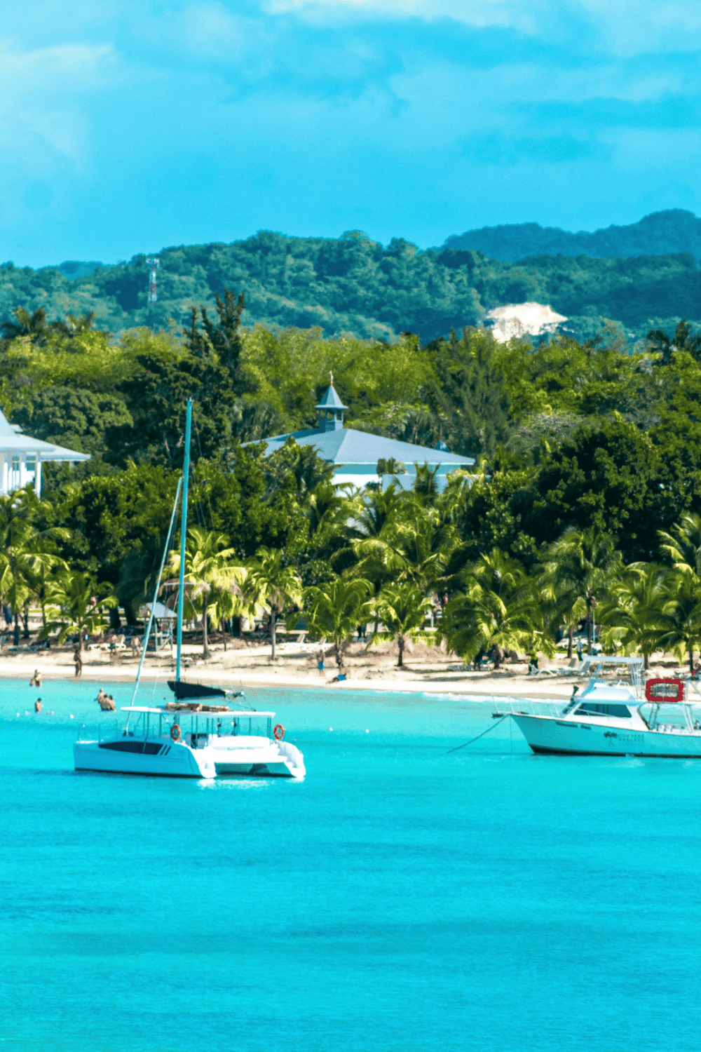Beach with sailboats and lush tropical trees under a blue sky.