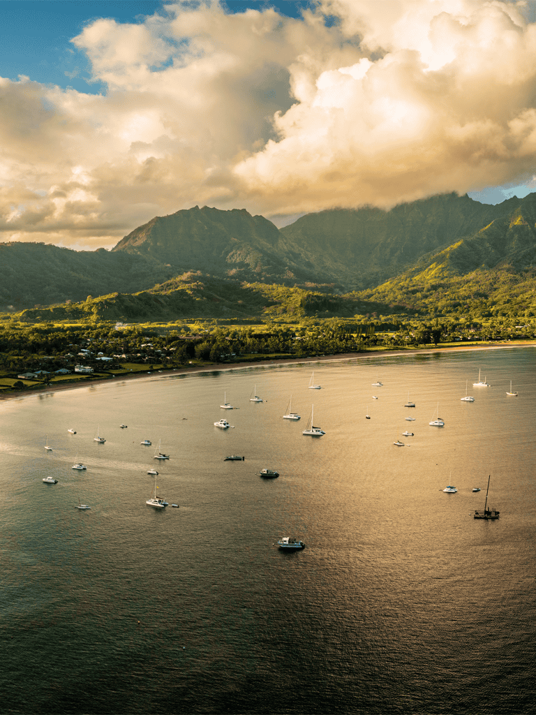 Remote mountain and harbor landscape with sailing boats in Hawaii, lush green hills, and dramatic cloud-filled sky, scenic and tranquil.