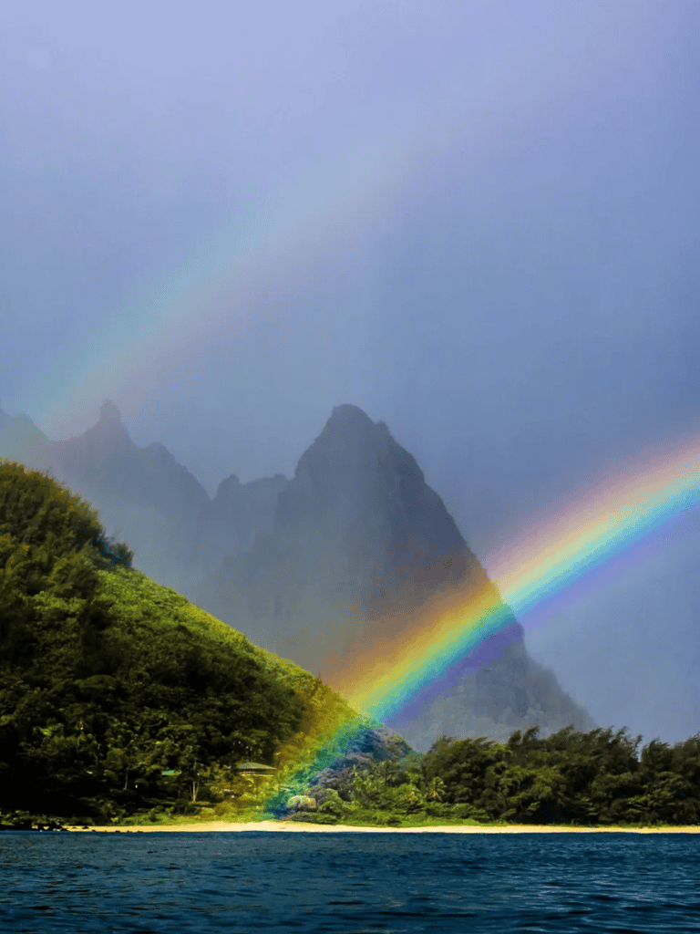 Colorful rainbow over lush green mountains and ocean, scenic natural landscape.