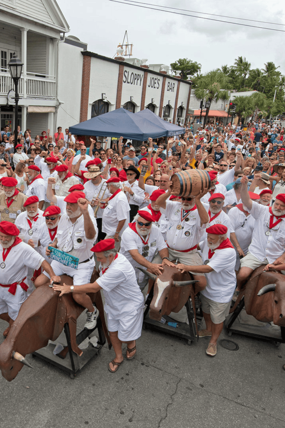 Crowd participating in a festival parade with bull sculptures and people wearing white shirts and red berets for an event in a lively town.