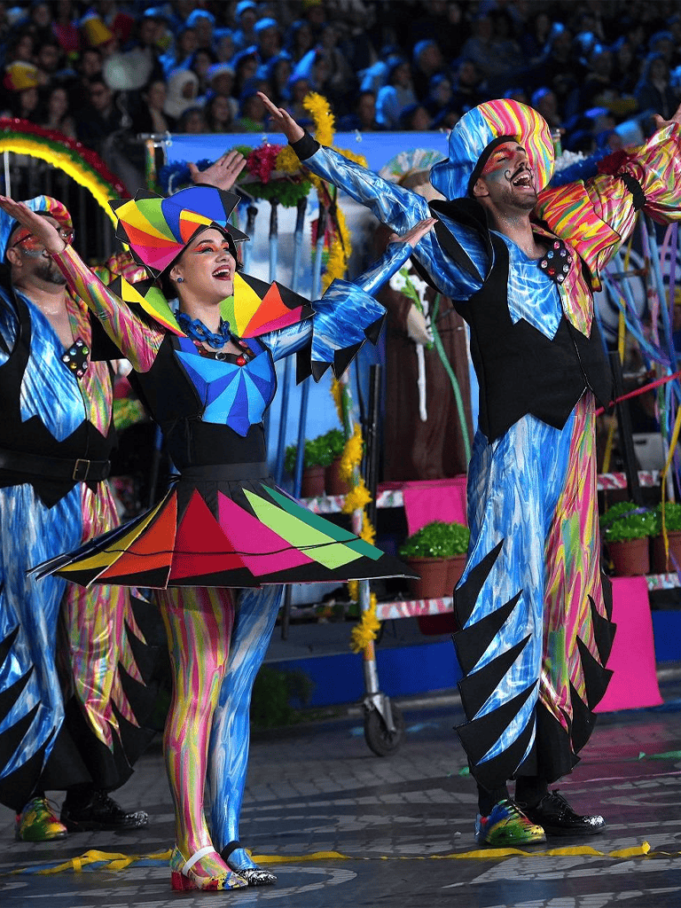 Vibrant performers in colorful costumes at a festive parade celebration, with a joyful crowd in the background.