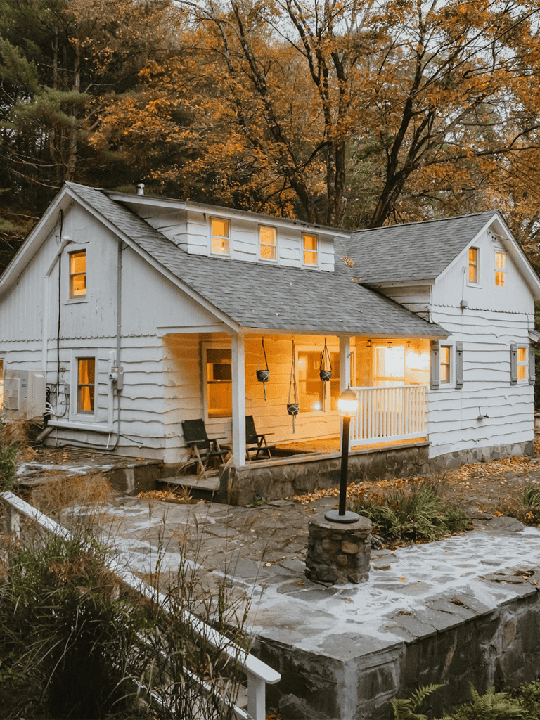 Cozy white cottage with warm lighting surrounded by autumn trees and outdoor seating.