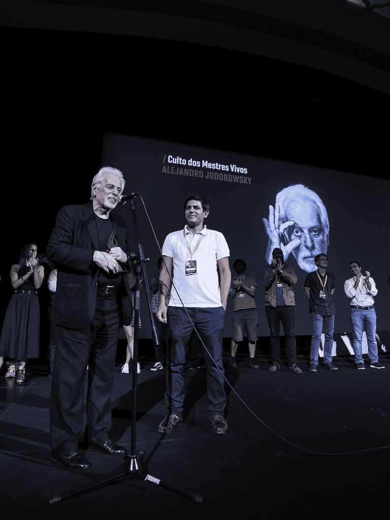 A man speaking at an event stage with a large screen displaying Alejandro Jodorowsky's image and name.