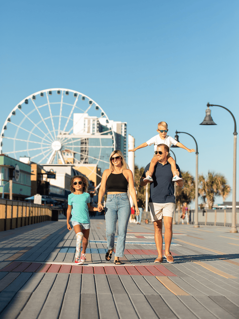 Family enjoying a day at a seaside amusement park with a Ferris wheel in the background.