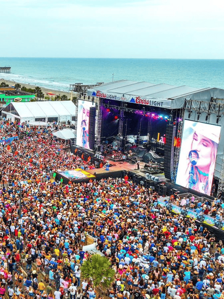 Vibrant beach concert with large crowd, stage, and ocean view for summer entertainment events.