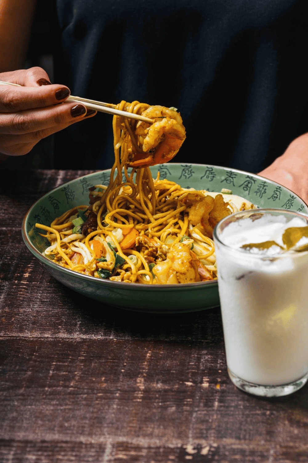 Savory Asian noodle dish with shrimp and vegetables on a wooden table, drink in the foreground.