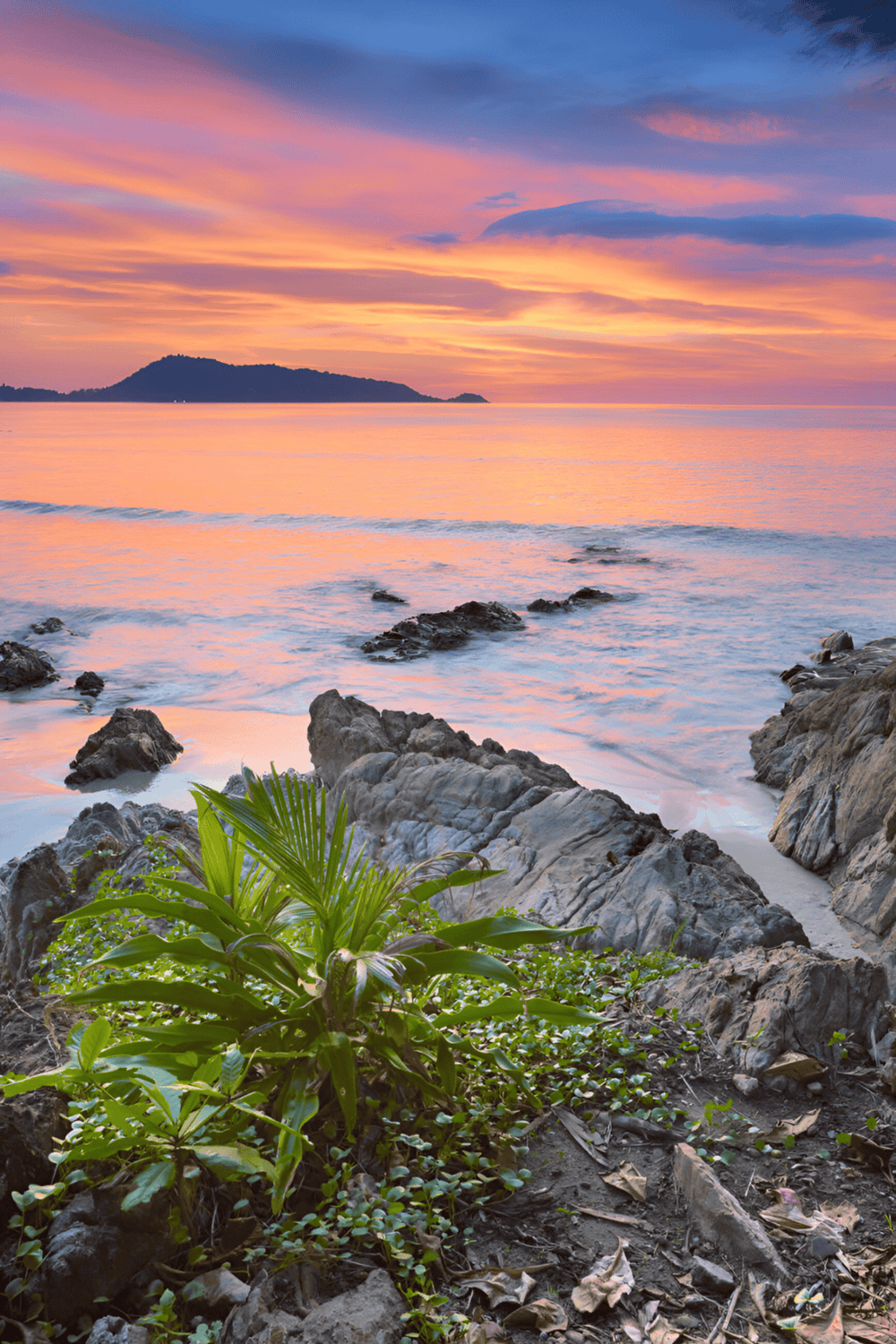 Vibrant sunrise over serene ocean with rocks and lush greenery on the shore, a perfect tropical getaway scene.
