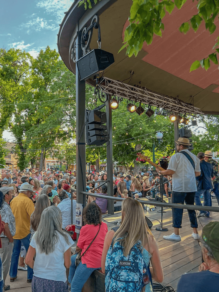 Crowd enjoying live outdoor music concert on stage in a park with lush green trees.
