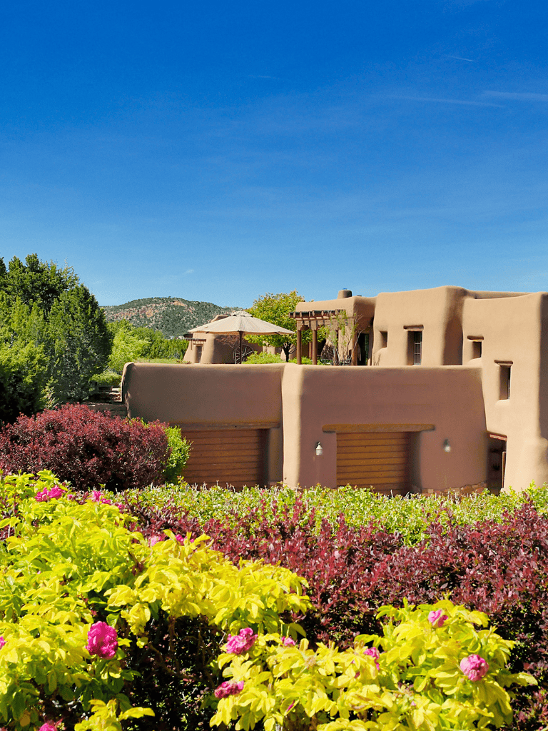 Adobe Southwestern-style house with lush garden and mountain view under bright blue sky.