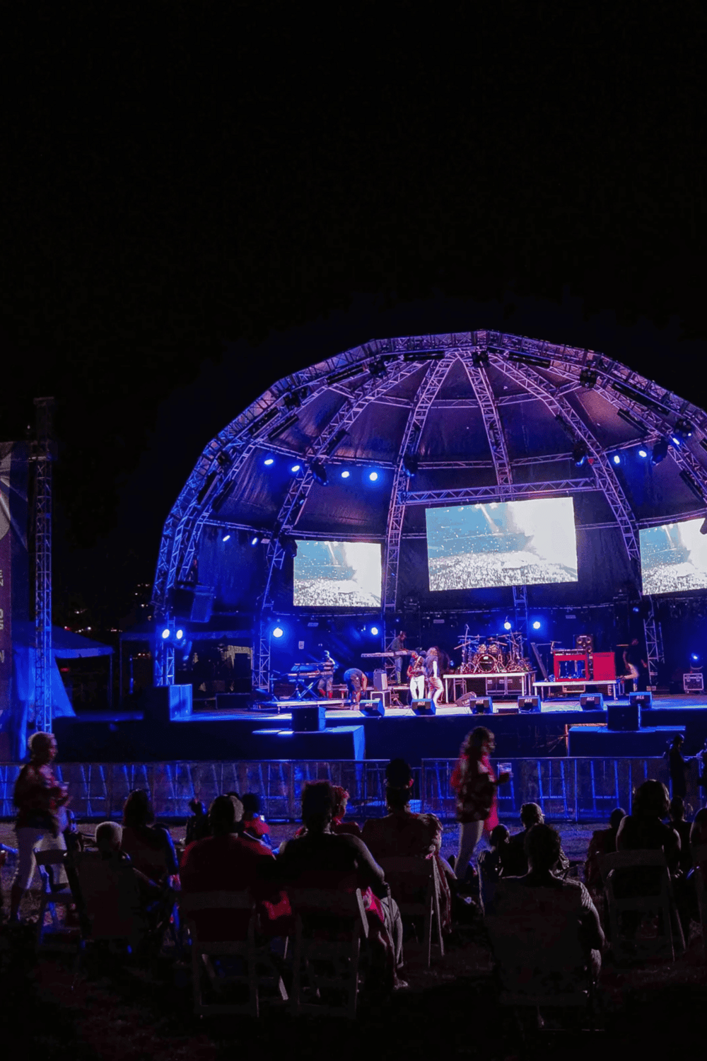 Outdoor concert stage at night with vibrant blue lighting and large screens, showcasing event entertainment and live music.