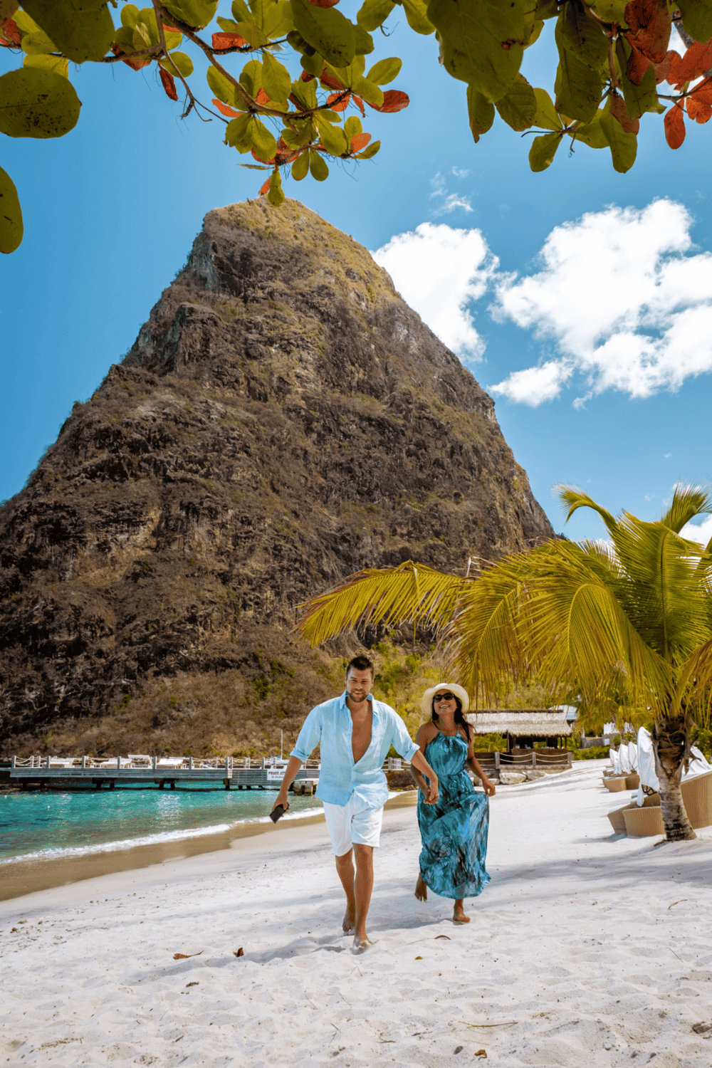 Vibrant beach scene with couple walking, lush palm trees, towering mountain, and clear blue sky, perfect for tropical travel and adventure.