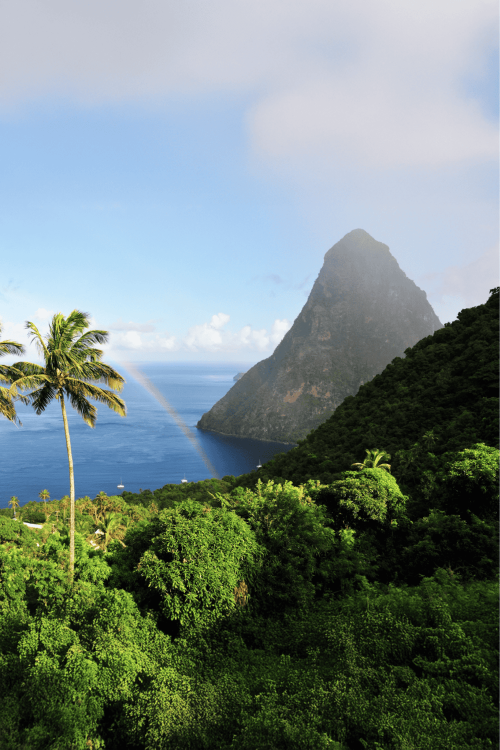 Vibrant tropical landscape with lush green trees, towering mountain, blue ocean, and rainbow on a sunny day.