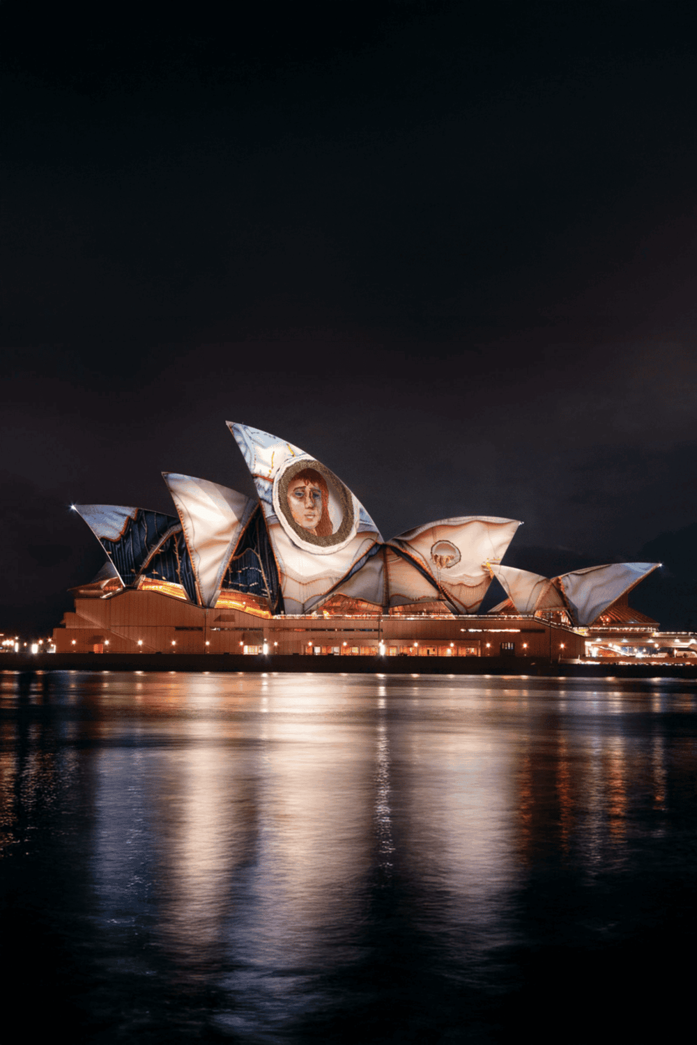 Sydney Opera House illuminated at night with artistic projections on its sails, reflecting on the water below.