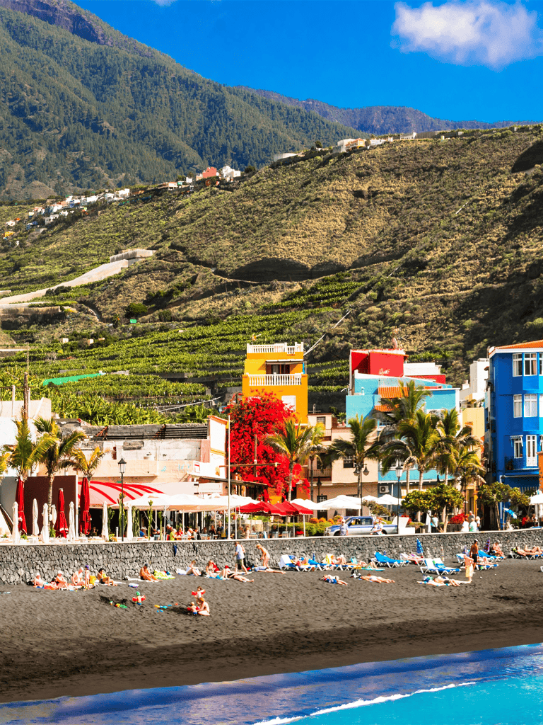 Serene beach in Puerto de la Cruz, with colorful buildings and lush green mountains in the background.