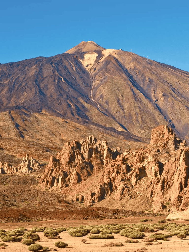 Vast volcanic mountain landscape with rugged rocks, desert terrain, and sparse vegetation under clear blue sky.