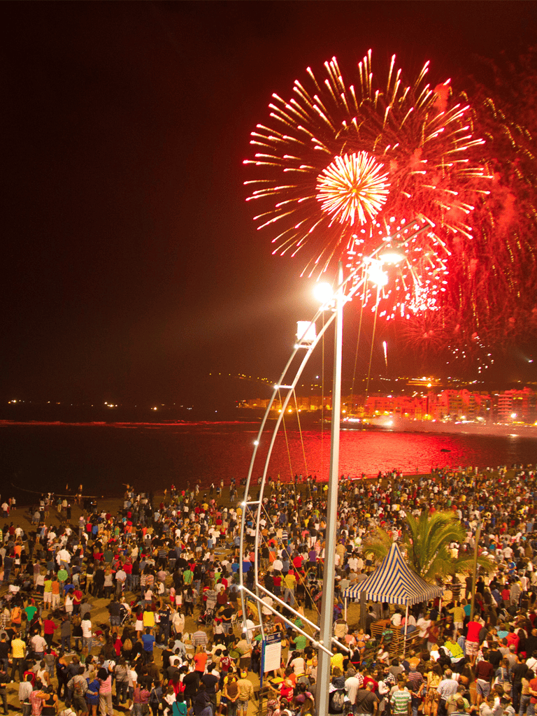 Bright fireworks over a crowded beach event celebrating a festive occasion, with a diverse crowd enjoying the fireworks show.