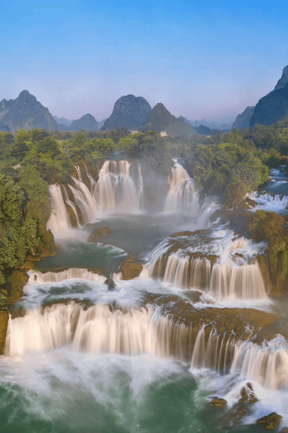 Vivid image of stunning waterfalls in lush green landscape with towering karst mountains in the background.