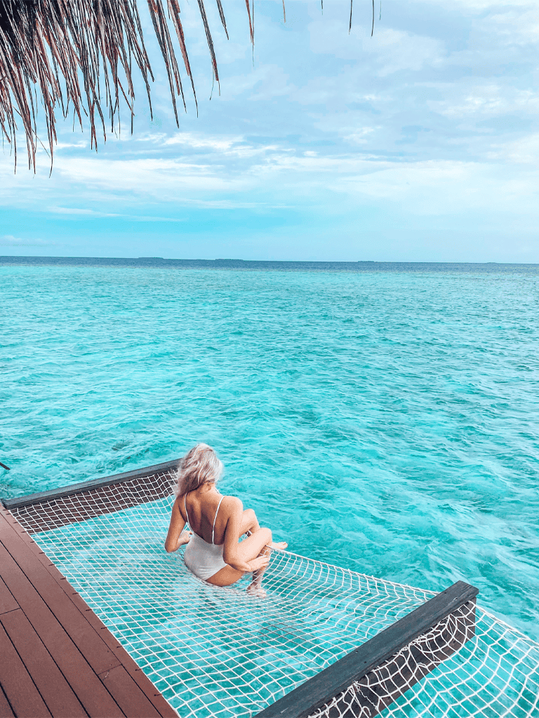 Relaxing on a tropical ocean overwater bungalow deck with clear blue water and palm thatching overhead.