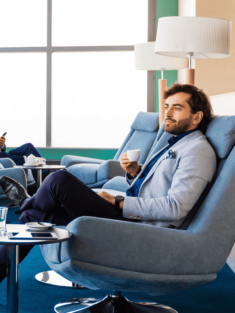 Relaxed businessman enjoying coffee in modern airport lounge with comfortable armchair and bright natural light.