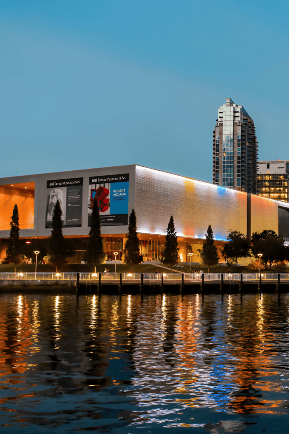 Stunning view of a modern arts center illuminated at dusk, reflecting on water, with city skyline in background.