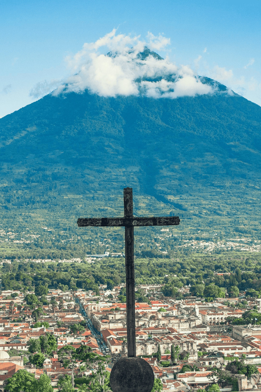 Panoramic view of a cross monument with an active volcano and cityscape in the background, highlighting travel and exploration destinations.