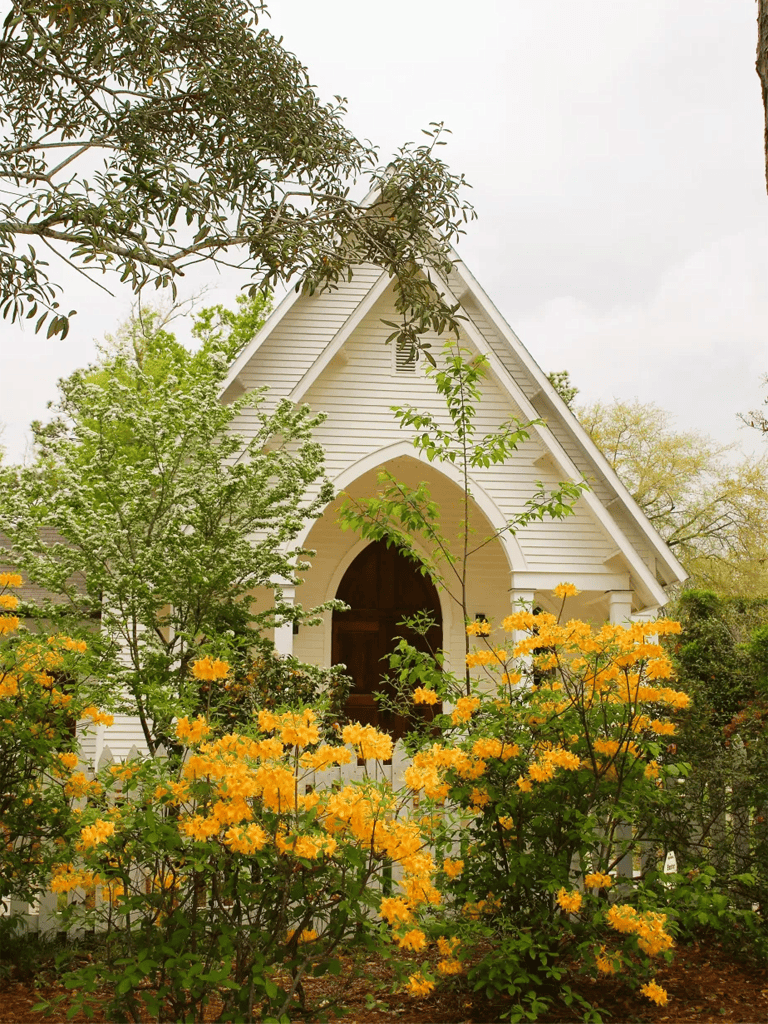 Chapel with white siding and yellow flowers in front, surrounded by trees, in a peaceful garden setting.
