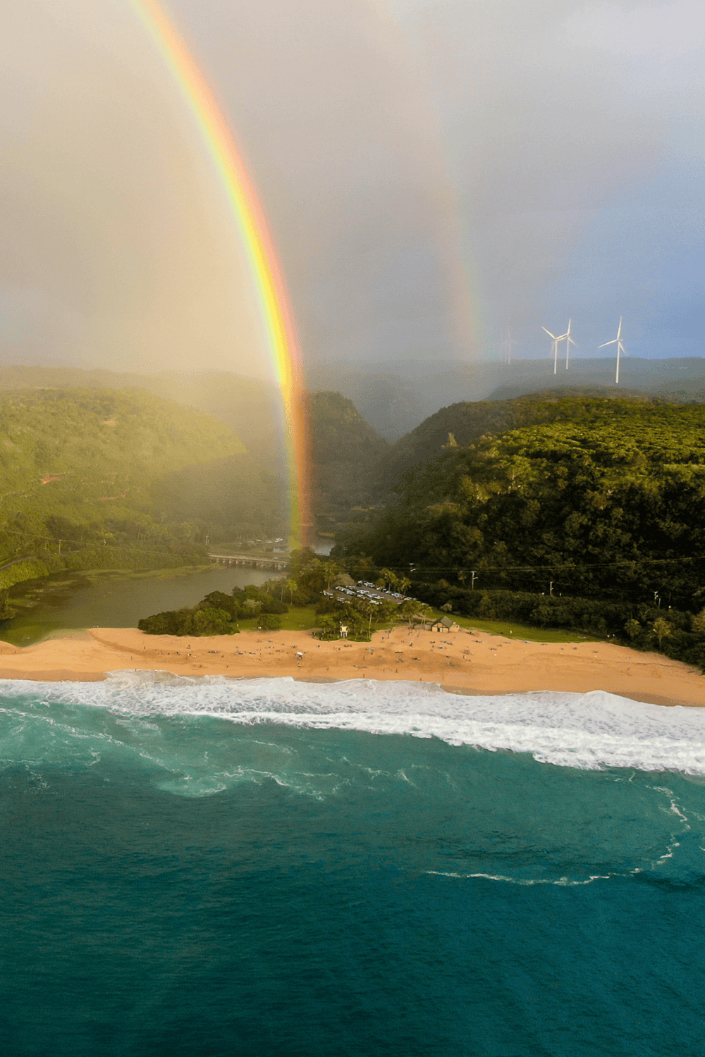 Vibrant rainbow over coastal green hills and wind turbines with ocean waves in foreground.