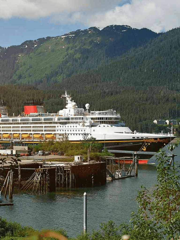Luxury cruise ship docked at scenic coastal port with lush green mountains in the background.