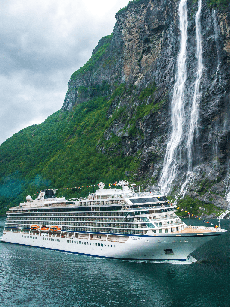 Luxurious cruise ship sailing near majestic waterfall in scenic fjord setting.