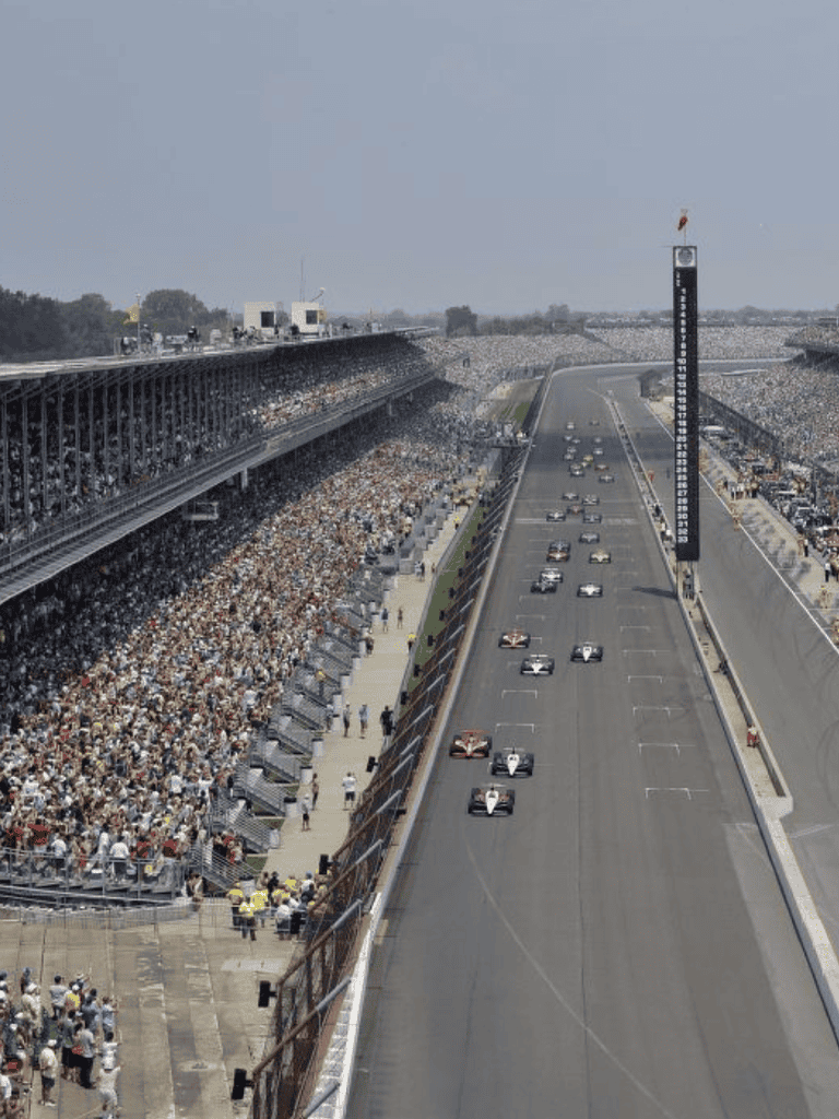 Huge crowd at a racing event with cars on the track and spectators in grandstands, bright sunny day, event venue.