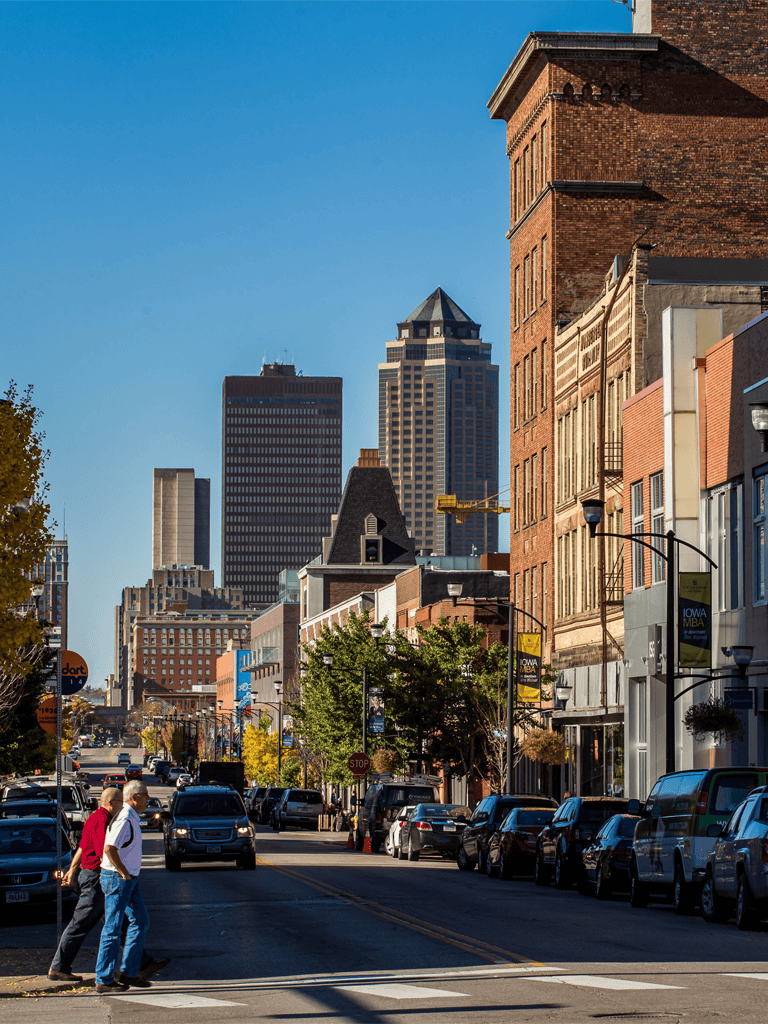 Wide city street with tall skyscrapers, historic buildings, and pedestrians walking on crosswalk.