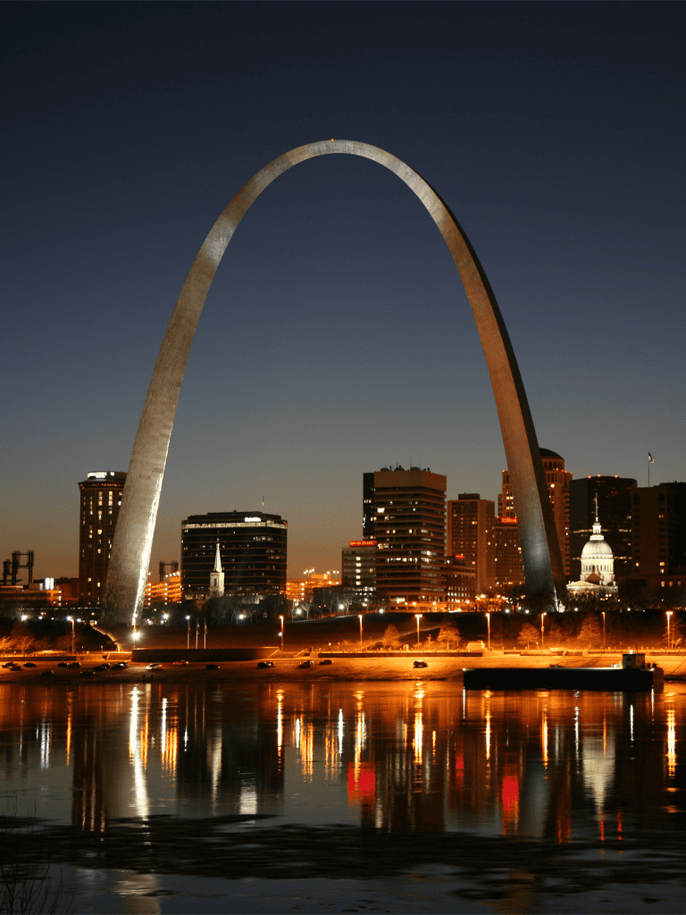 Arch of St. Louis at night in downtown St. Louis with illuminated cityscape reflection on water.