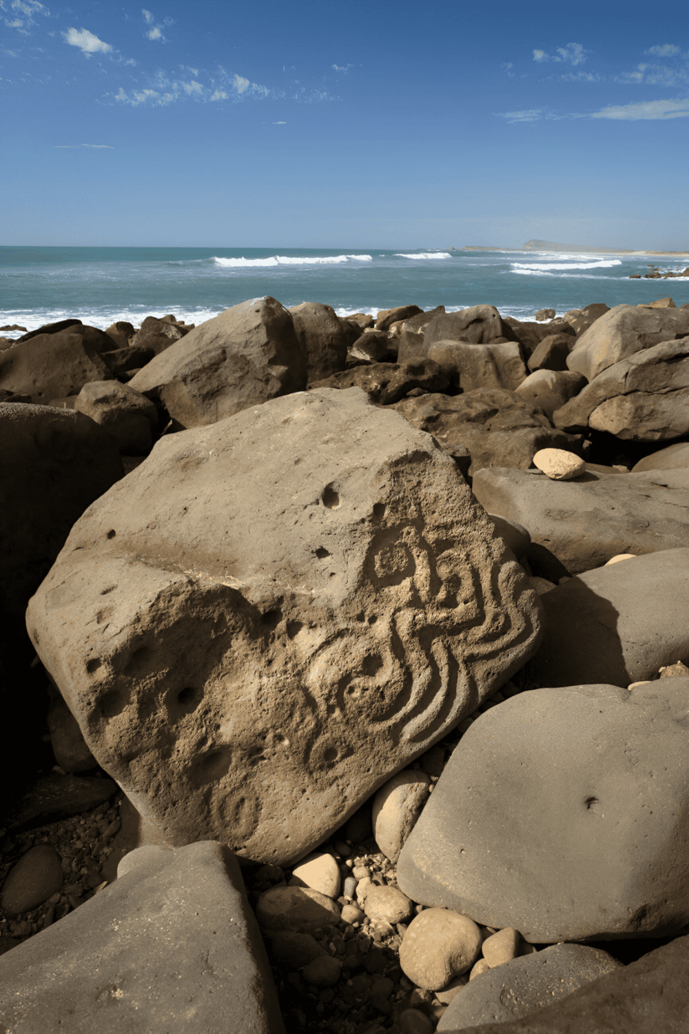 Ancient rock carving along the rugged coastline with ocean waves and clear blue sky in the background.