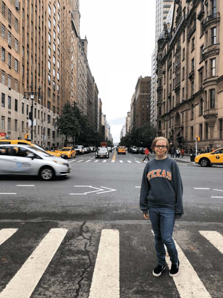 Girl standing at crosswalk on a city street filled with taxis and cars, urban architecture in the background, QuestForDirections.