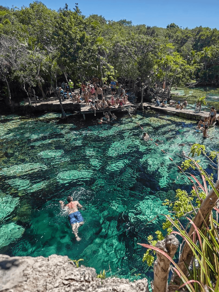 Brightly colored outdoor swimming hole surrounded by lush green trees, with swimmers enjoying the clear water and bright sunshine.