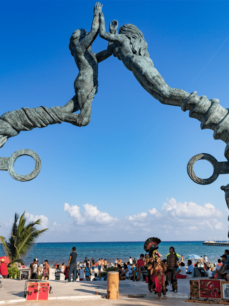High-rise statue of two intertwined figures high-fiving on a beach with visitors, palm trees, and ocean in the background, showcasing cultural art and outdoor scene.