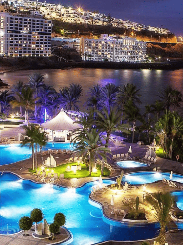 Luxurious nighttime resort pool area with palm trees and cityscape skyline in the background.