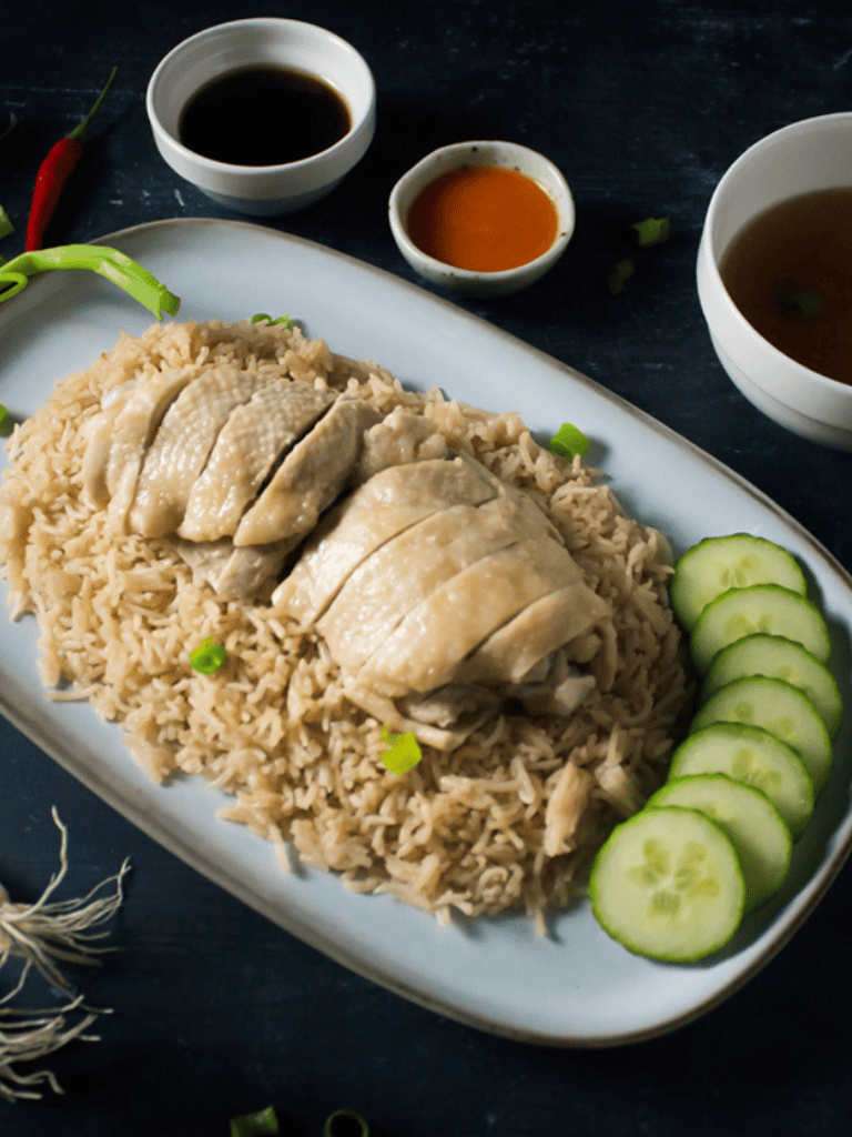 Poached chicken with jasmine rice, fresh cucumber slices, soy sauce, and hot sauce on a white platter.